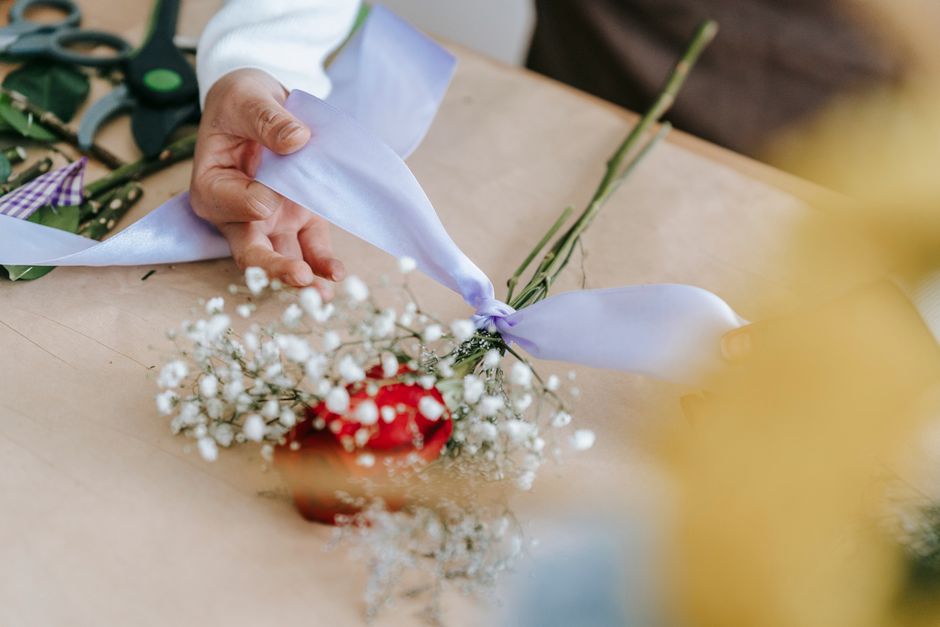 A florist's hand arranging a floral gift featuring a small bouquet of white baby's breath and a single red flower, wrapped with a purple ribbon, on a light wooden surface. The arrangement is in the pr
