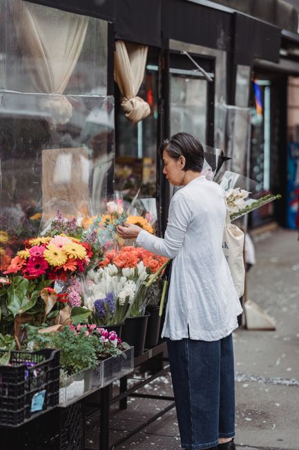 A woman with short dark hair, dressed in a light grey cardigan and dark jeans, selecting a floral arrangement from a display outside a flower shop on Eltham High Street. The bouquet features a mix of