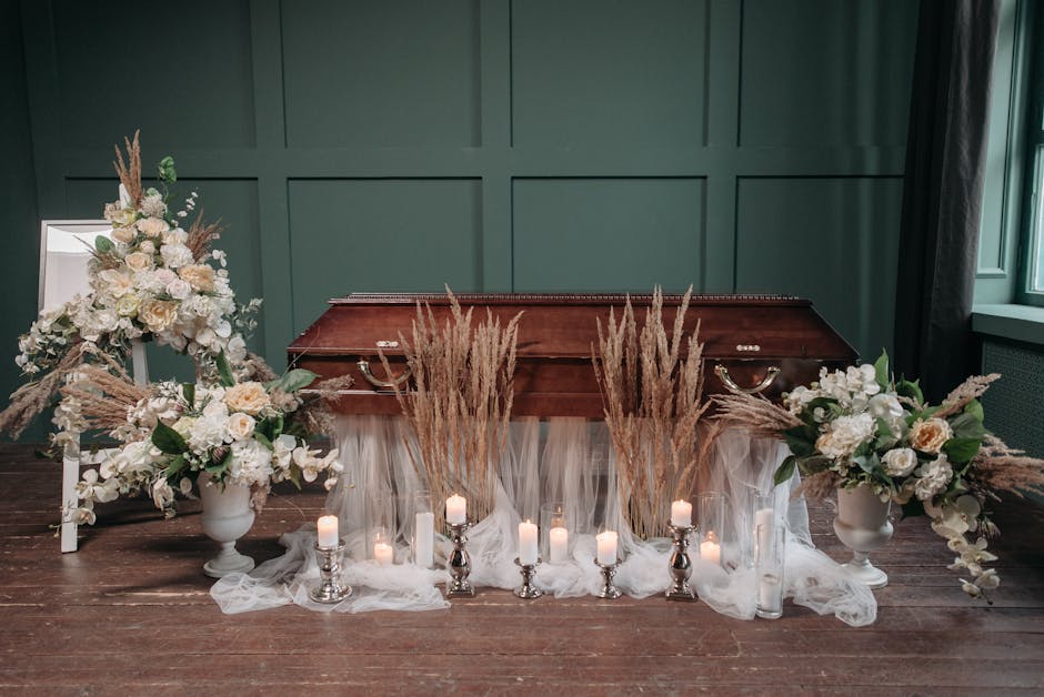 A funeral floral arrangement placed on a wooden floor against a green paneled wall. The display features two large white urns filled with pale pink and white roses, white lilies, and greenery, accente