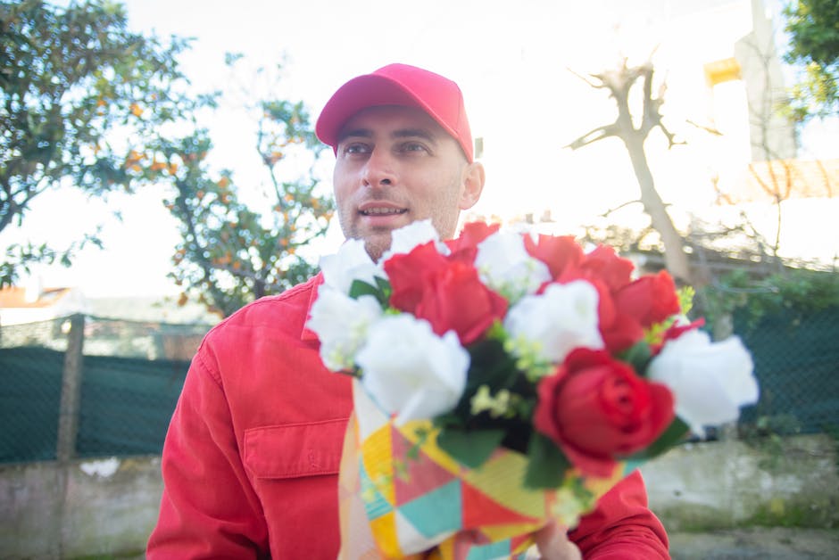 A male flower delivery person dressed in a red uniform and matching cap is holding a vibrant bouquet of fresh flowers, consisting of red and white roses and other blooms, wrapped in colorful paper. Th