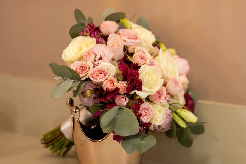 A bouquet of fresh flowers arranged in a rounded, dense style features predominantly pale pink and white roses, complemented by deep burgundy flowers and sprigs of light green eucalyptus leaves. The b